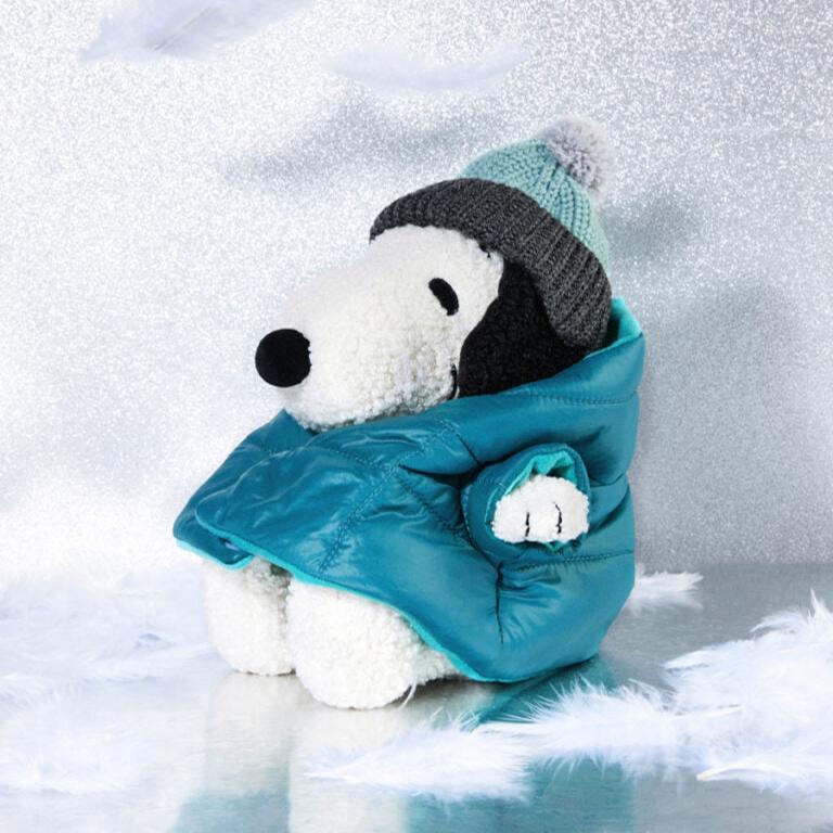 Stuffed Snoopy toy wearing a teal puffer coat and gray and mint knit hat against a glittery background.
