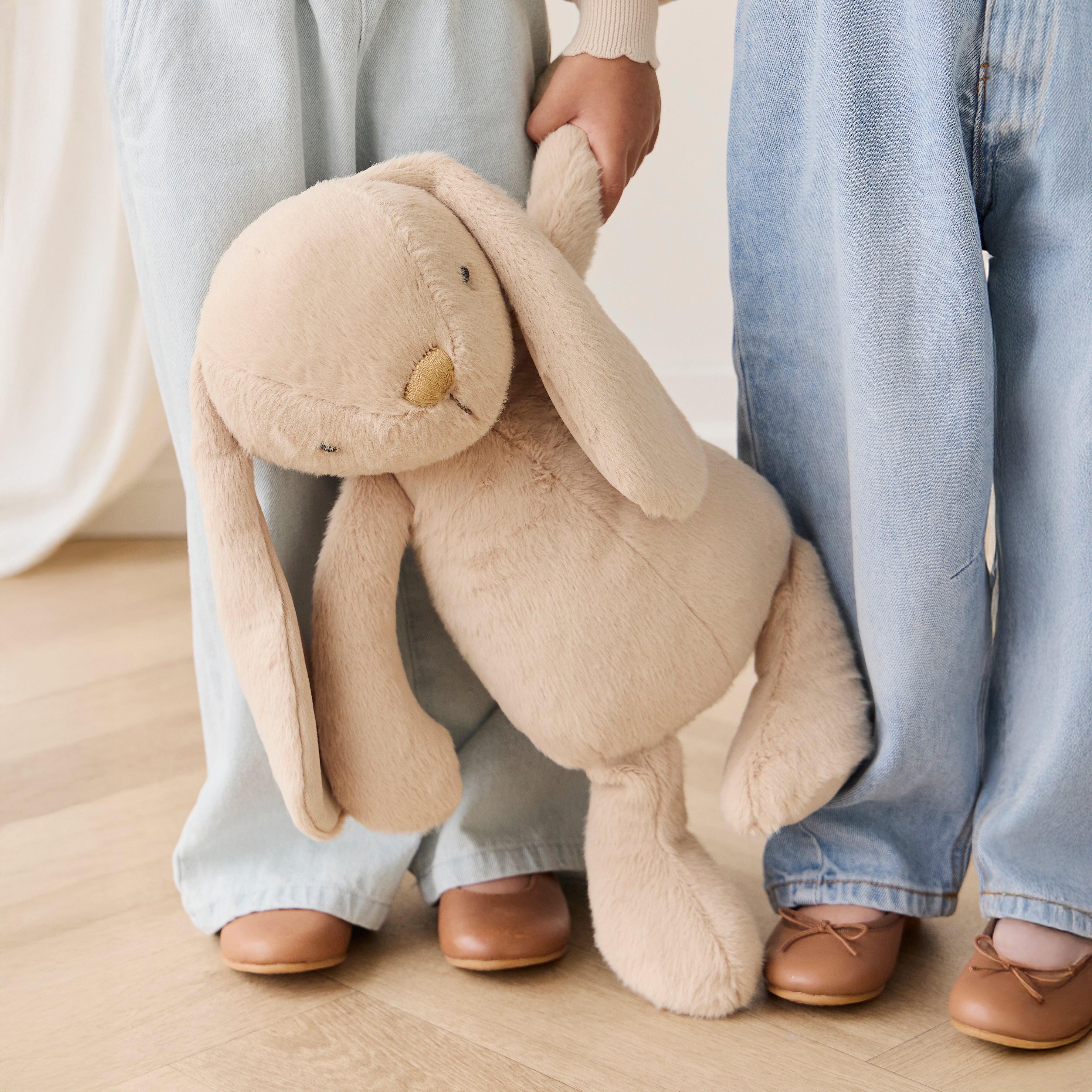 Two people holding a large beige plush bunny toy on a wooden floor.