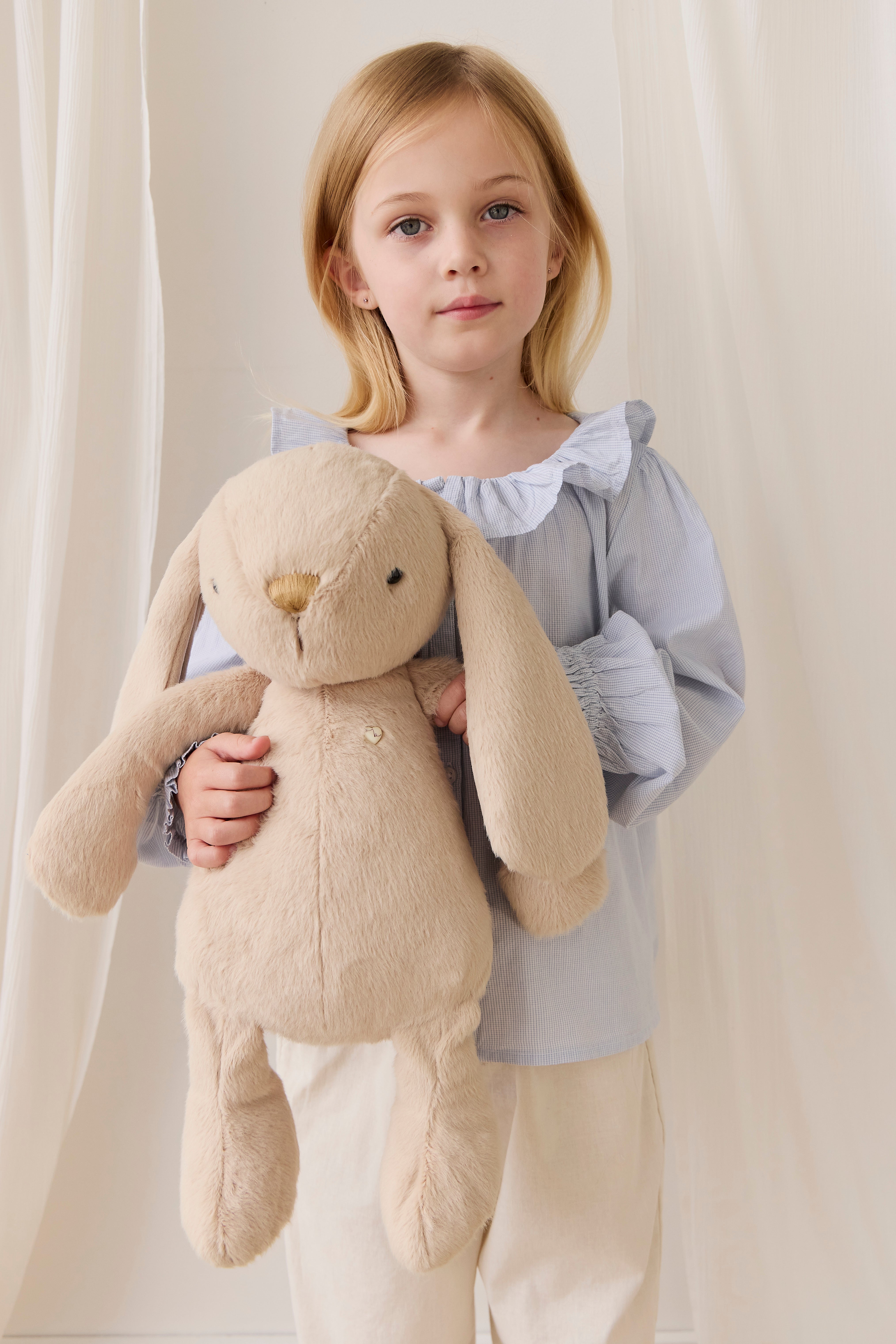 Young girl holding a beige stuffed bunny against a white curtain background