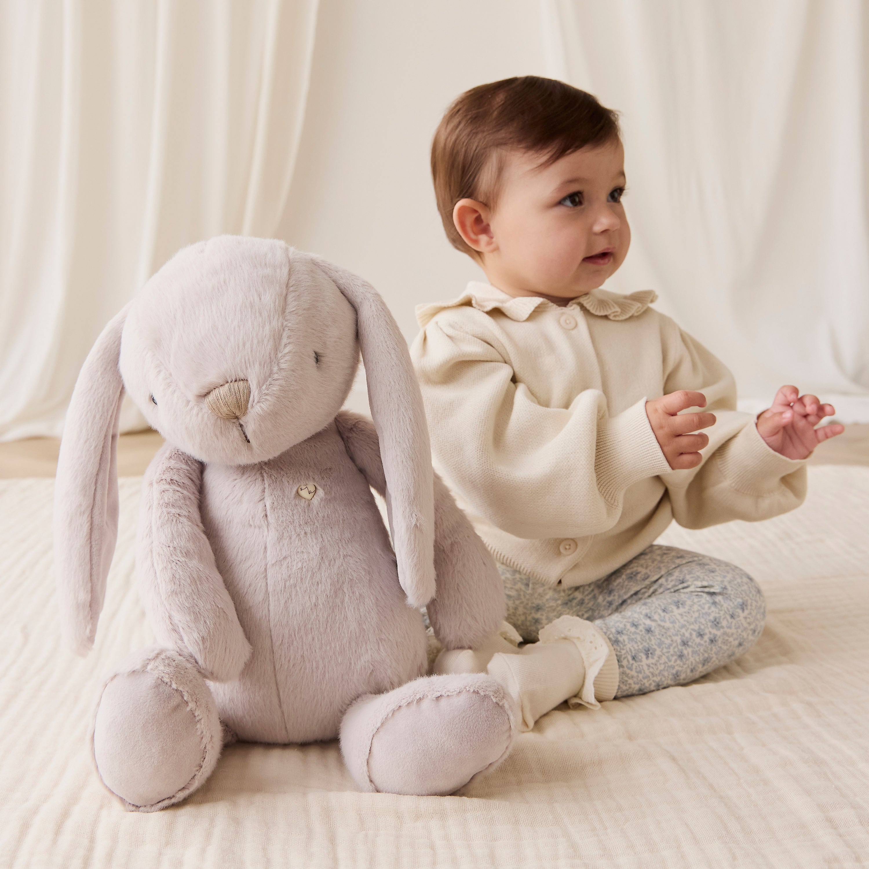 Baby sitting next to a plush rabbit toy on a soft surface