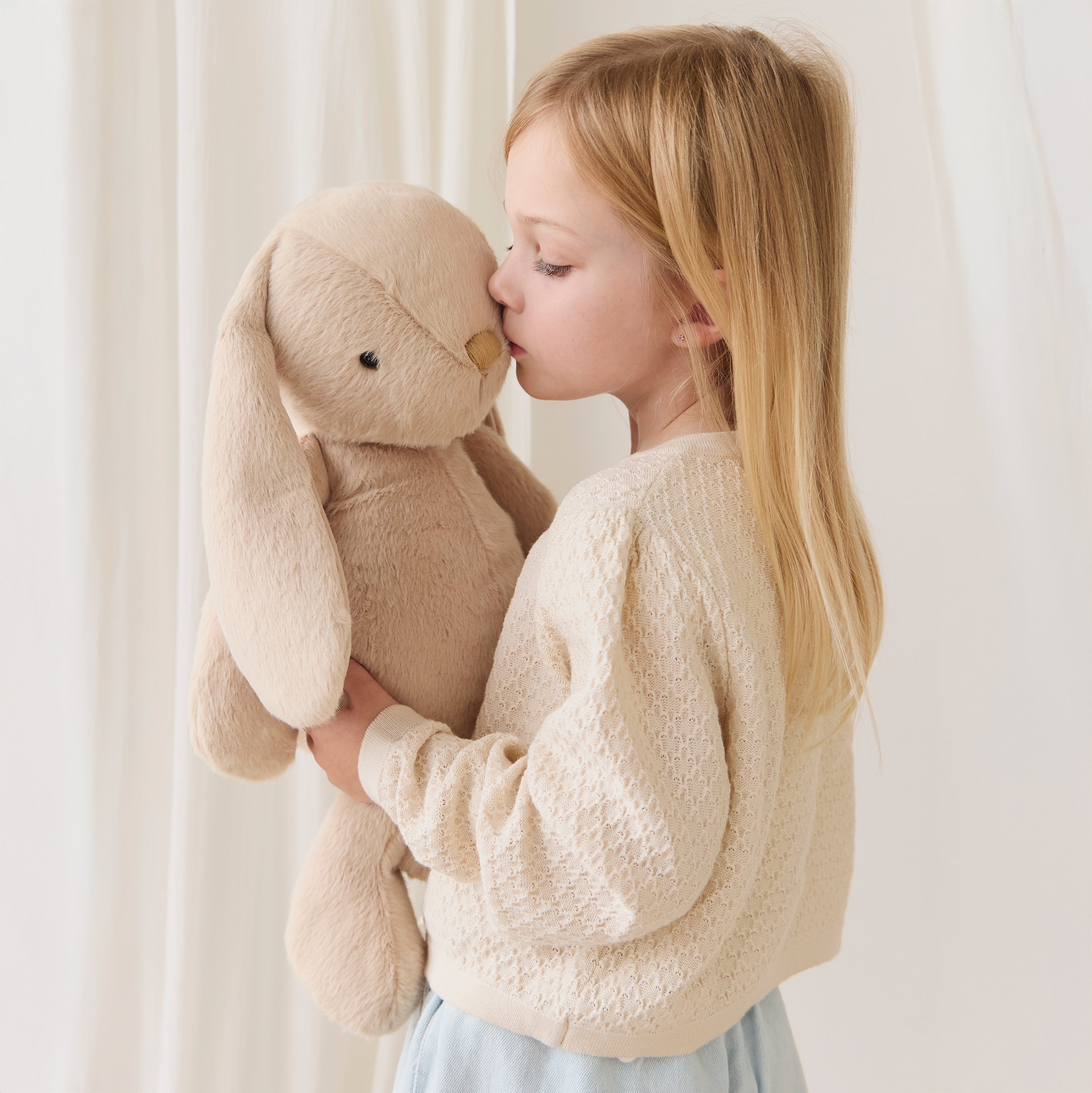 Young girl kissing a beige stuffed bunny toy against a white curtain background