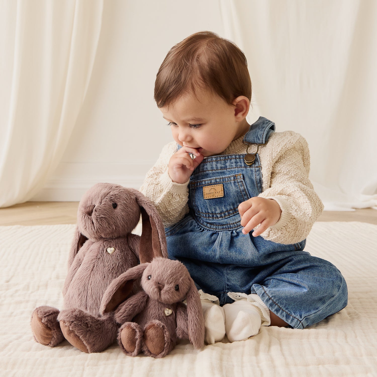 Child in denim overalls sitting with plush bunny toys on a soft surface
