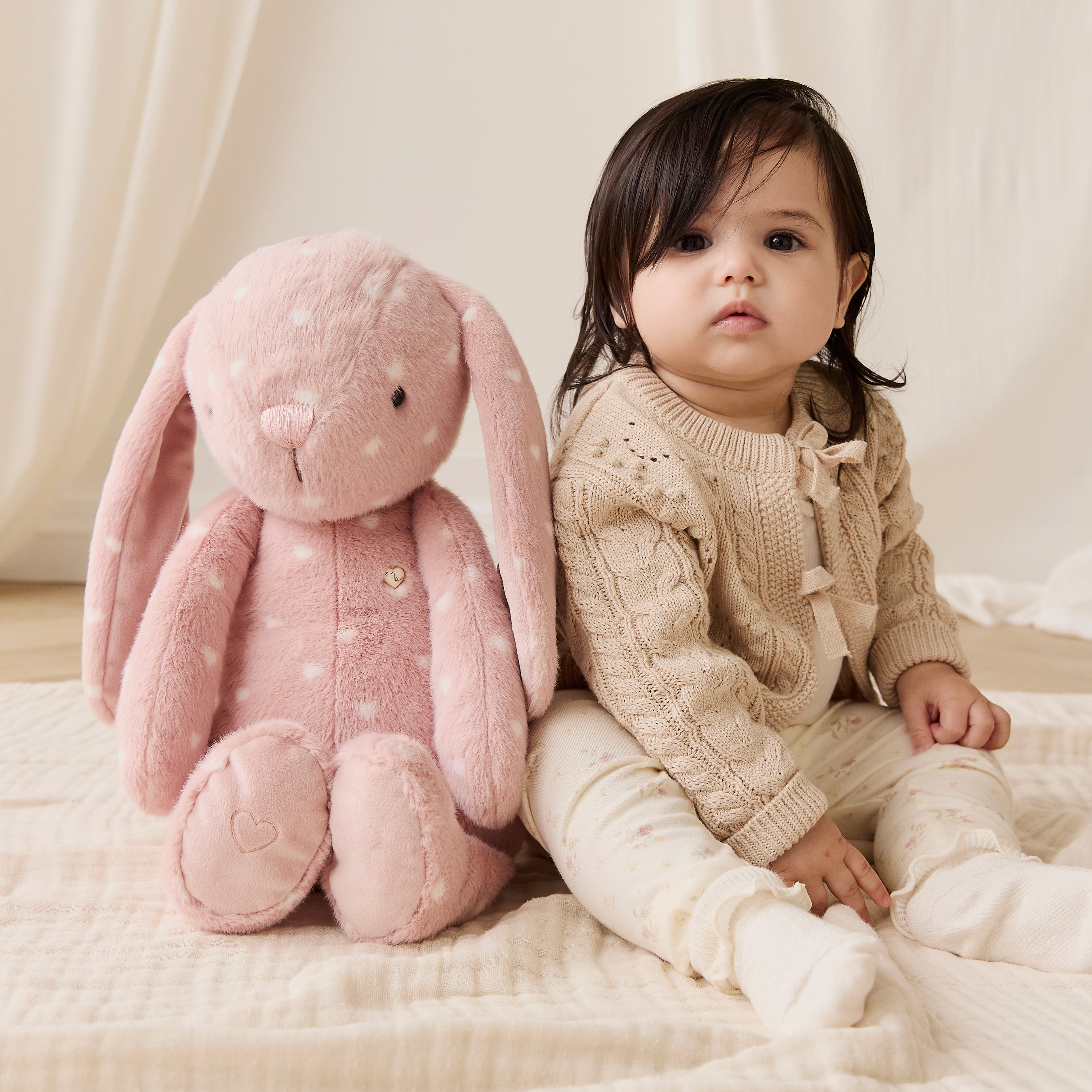 Child sitting next to a pink plush bunny toy on a soft surface