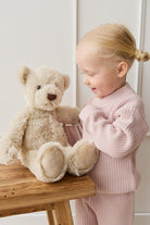 Girl in pink knitwear playing with teddy bear on wooden stool.