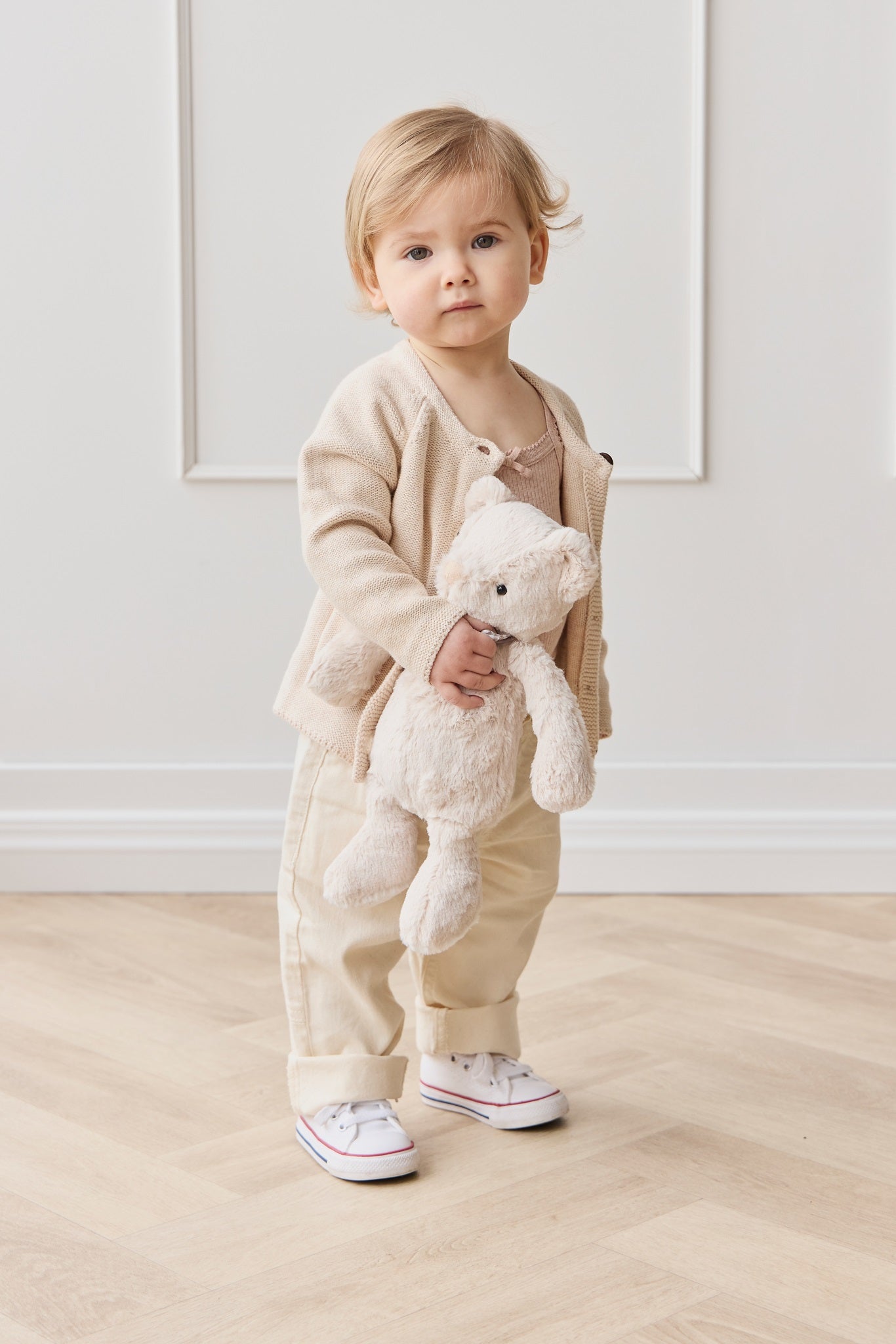 Full shot of a blonde toddler holding a teddy bear and standing in front of a white wall wearing a beige cardigan, pants, and white sneakers.