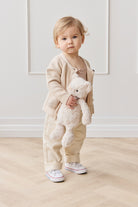 Full shot of a blonde toddler holding a teddy bear and standing in front of a white wall wearing a beige cardigan, pants, and white sneakers.