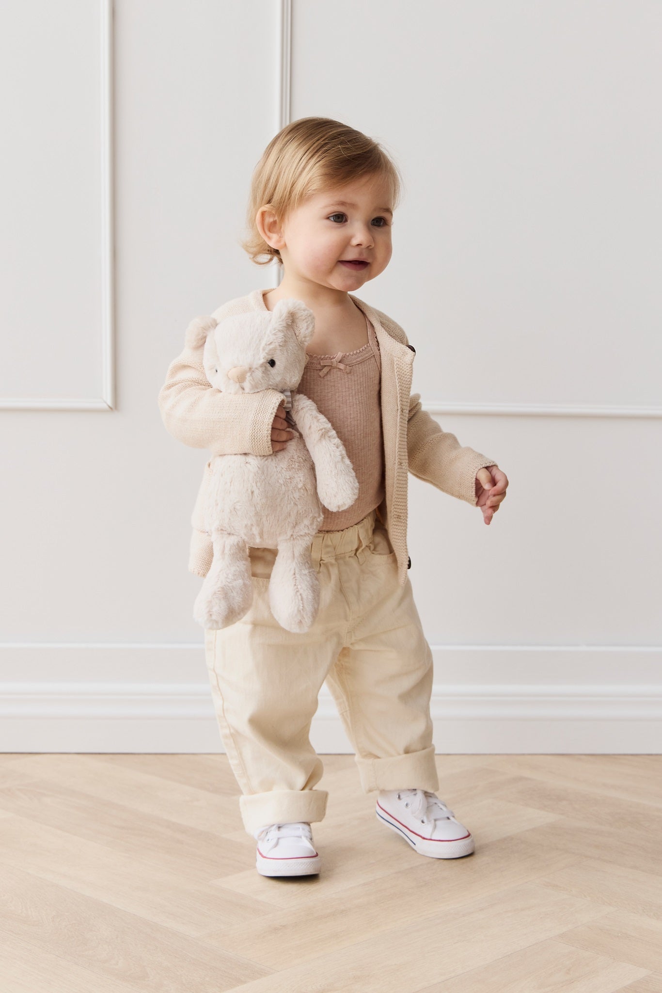 A toddler stands indoors, holding a teddy bear, wearing beige clothing and white sneakers.