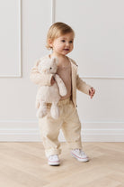 A toddler stands indoors, holding a teddy bear, wearing beige clothing and white sneakers.