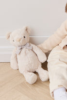Stuffed beige teddy bear with striped bow, sitting next to child in cream sweater and pants.