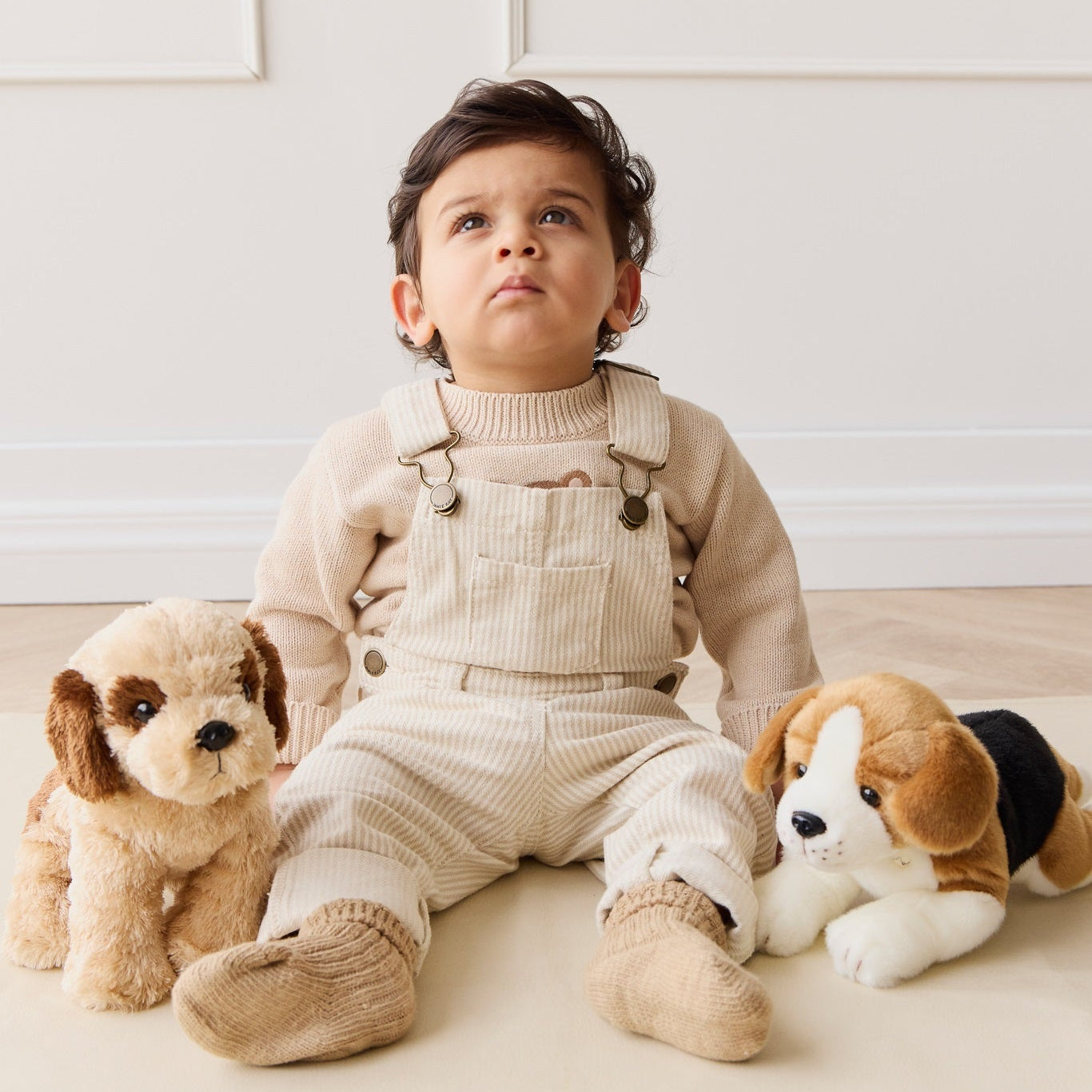 Infant sitting with plush dogs, wearing beige sweater, overalls, and booties.