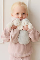 Blond toddler holding two plush bunnies, wearing a pink knitted outfit, against a white wall.