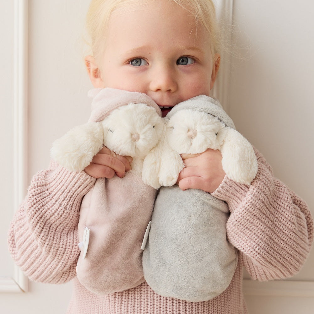 Blond toddler holding two plush bunnies, wearing a pink knitted outfit, against a white wall.