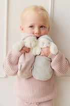 Blonde girl holding two plush bunny toys. She wears a pink knit outfit and stands against a white wall.