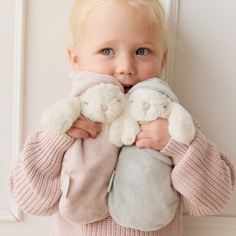 Blonde girl holding two plush bunny toys. She wears a pink knit outfit and stands against a white wall.