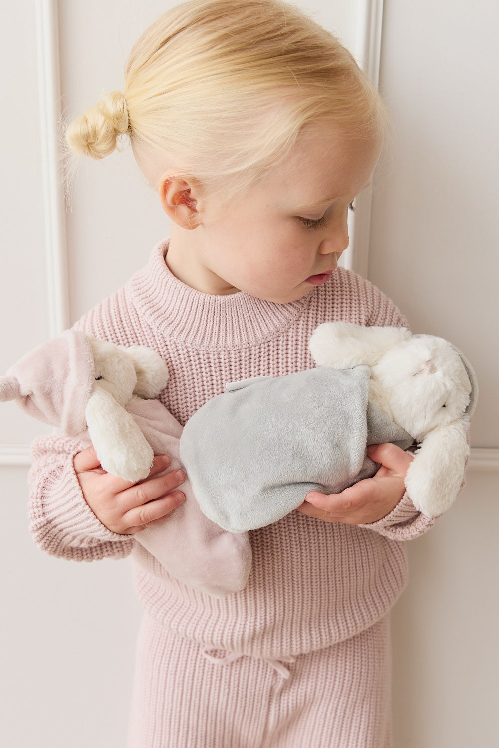 Blonde child holding two plush toys in pink knitwear.
