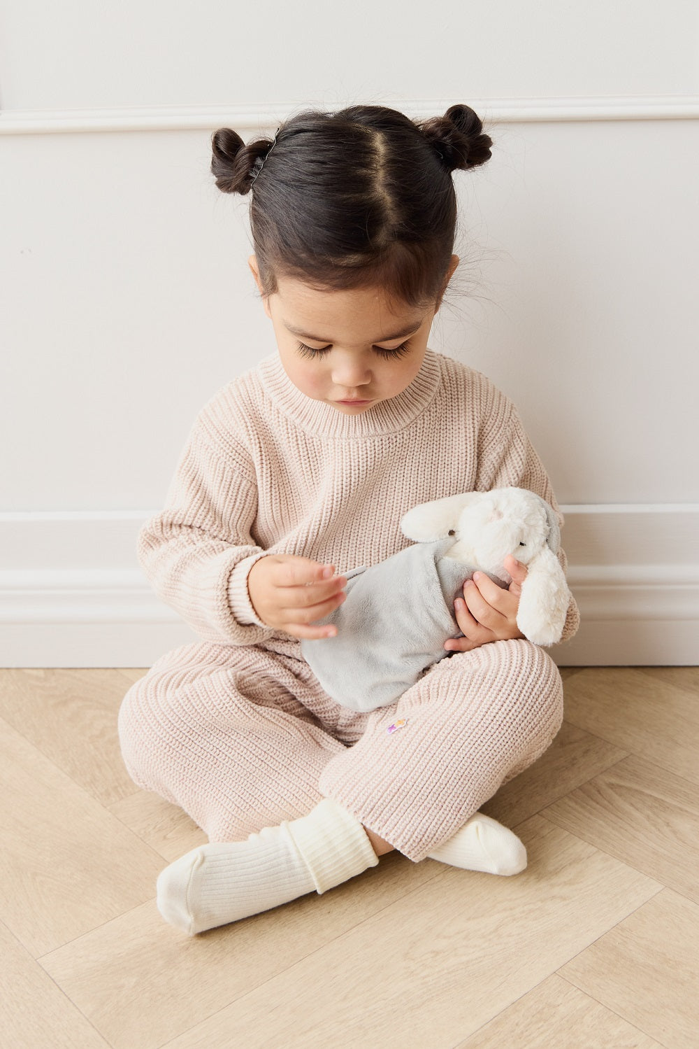 Toddler girl in beige knit outfit with white bunny stuffed animal, sitting on the floor.