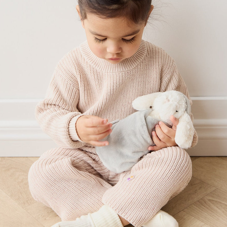 Toddler girl in beige knit outfit with white bunny stuffed animal, sitting on the floor.