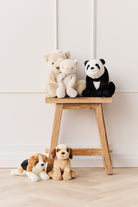 Stuffed teddy bears and dogs displayed on and around a wooden stool against a white wall and wooden floor.