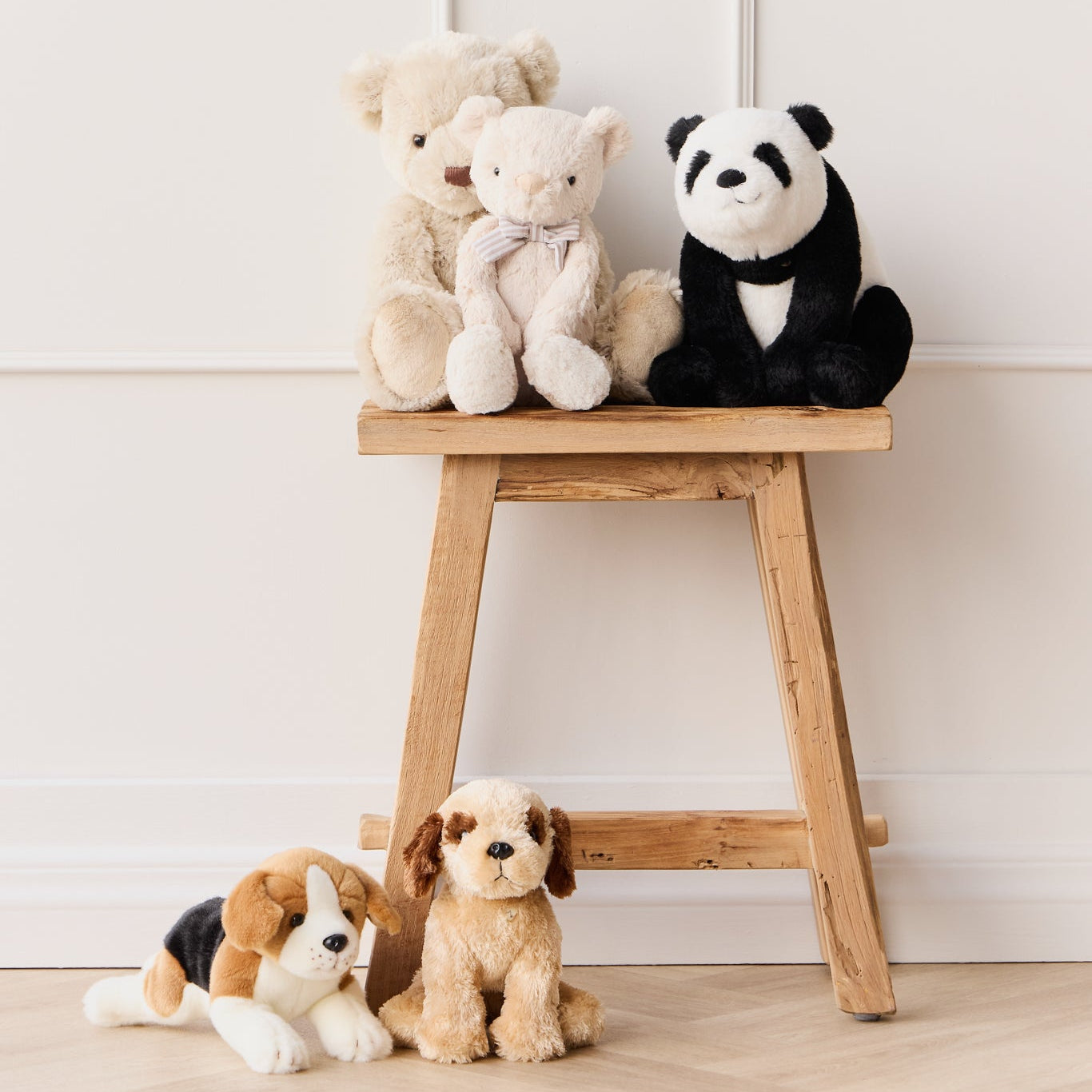 Stuffed teddy bears and dogs displayed on and around a wooden stool against a white wall and wooden floor.
