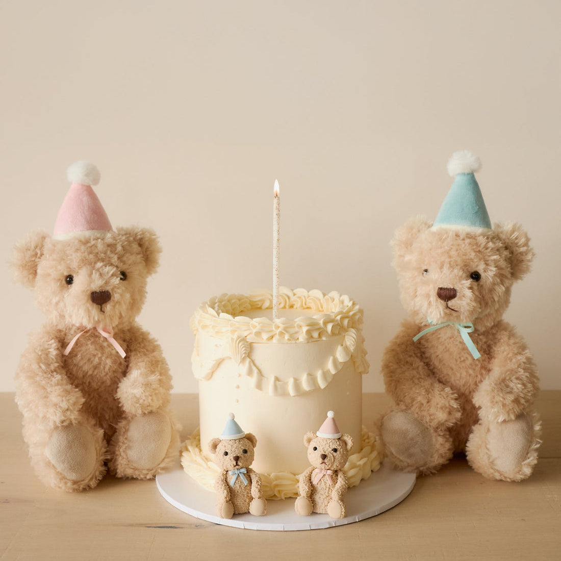 Two teddy bears wearing party hats flanking a cake with a lit candle, with two smaller teddy bear figurines in front of the cake.
