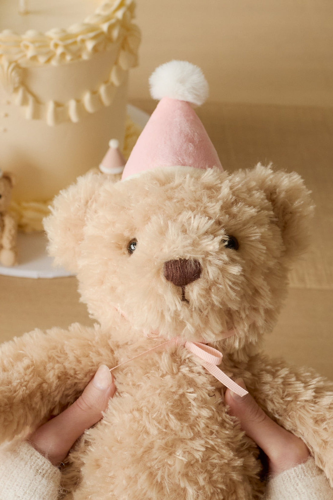 Close-up of a tan teddy bear wearing a pink party hat with white pompom, held by hands in a cream sweater, with a cake in the background.