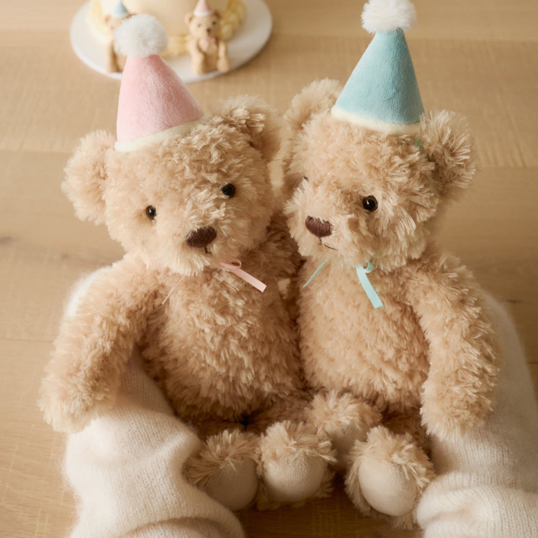 Two plush teddy bears wearing party hats are nestled in someone's arms.