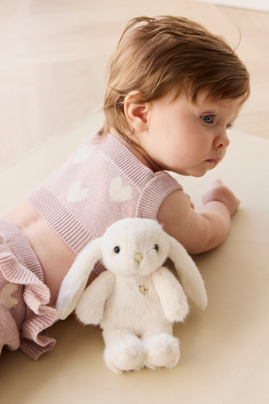 Baby lying with plush bunny toy, wearing pink knit top with white hearts.