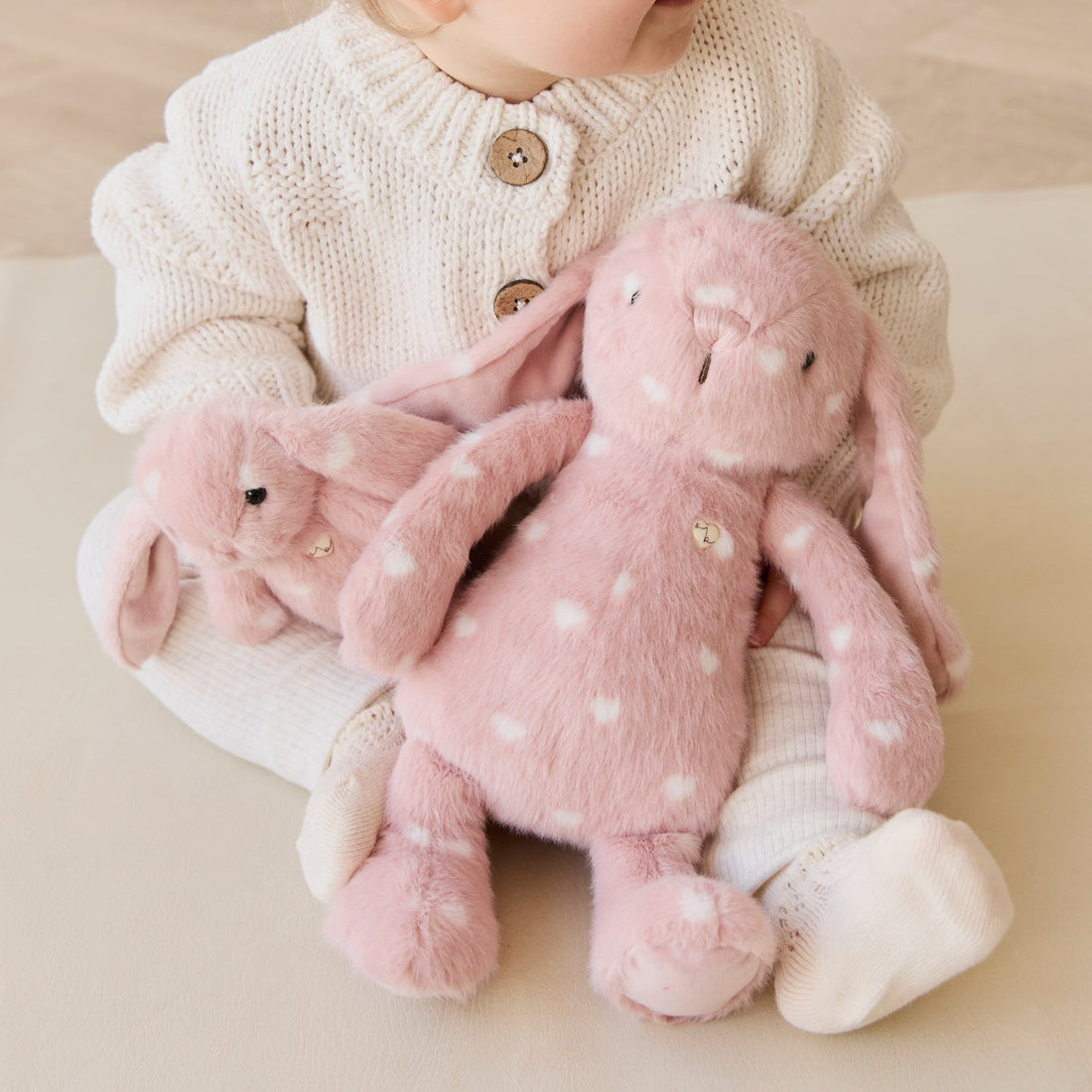 A child wearing a white knit sweater and white leggings sits holding two pink plush bunny toys with white polka dots.