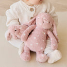 A child wearing a white knit sweater and white leggings sits holding two pink plush bunny toys with white polka dots.