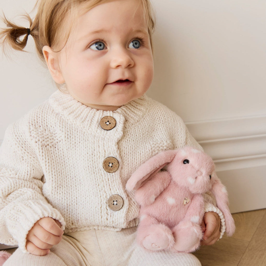 Baby in cream-colored knit cardigan with wooden buttons, holding pink plush bunny.