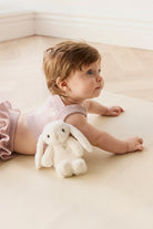 Baby lying on tummy with stuffed bunny, wearing pink knit romper on beige surface.