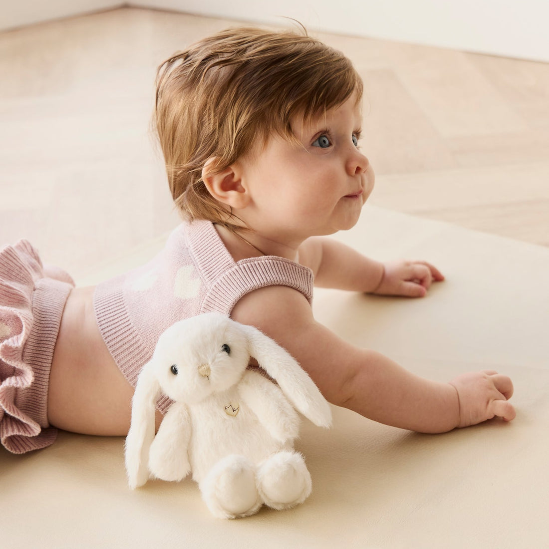 Baby lying on tummy with stuffed bunny, wearing pink knit romper on beige surface.