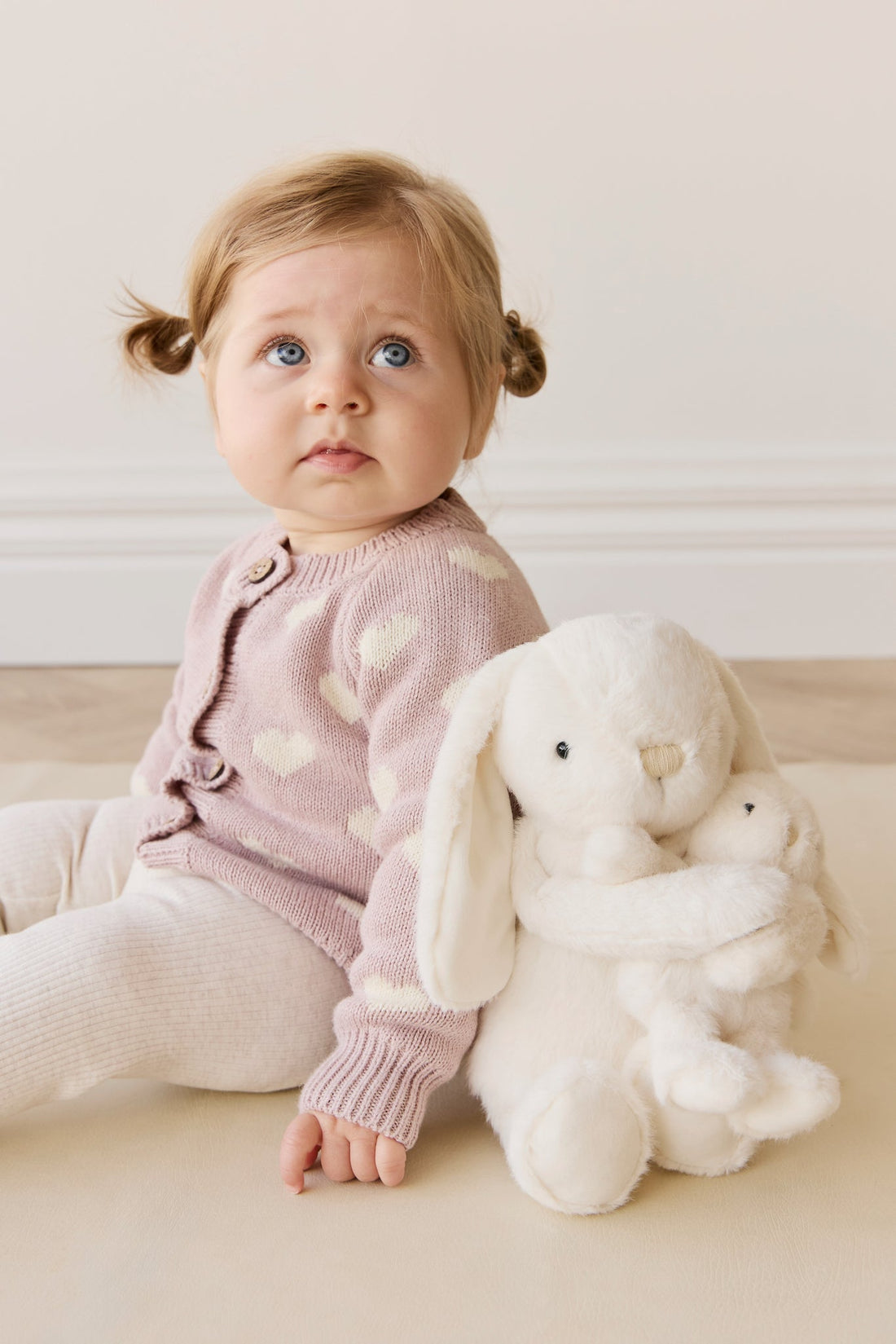 Child in pink sweater with heart patterns sitting on a light-colored floor with a white plush toy.