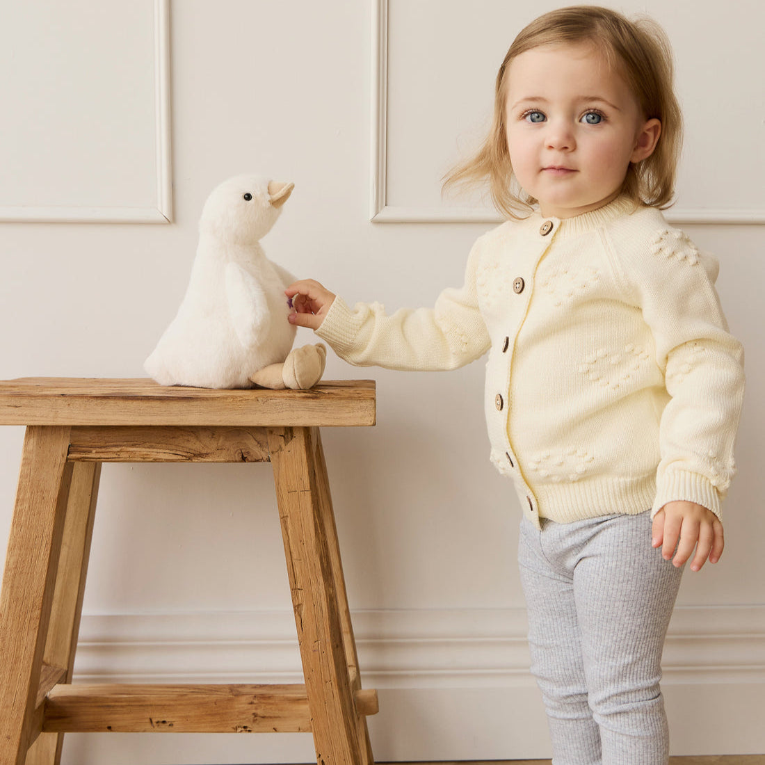 Toddler girl in yellow cardigan with stuffed white duck on wooden stool.