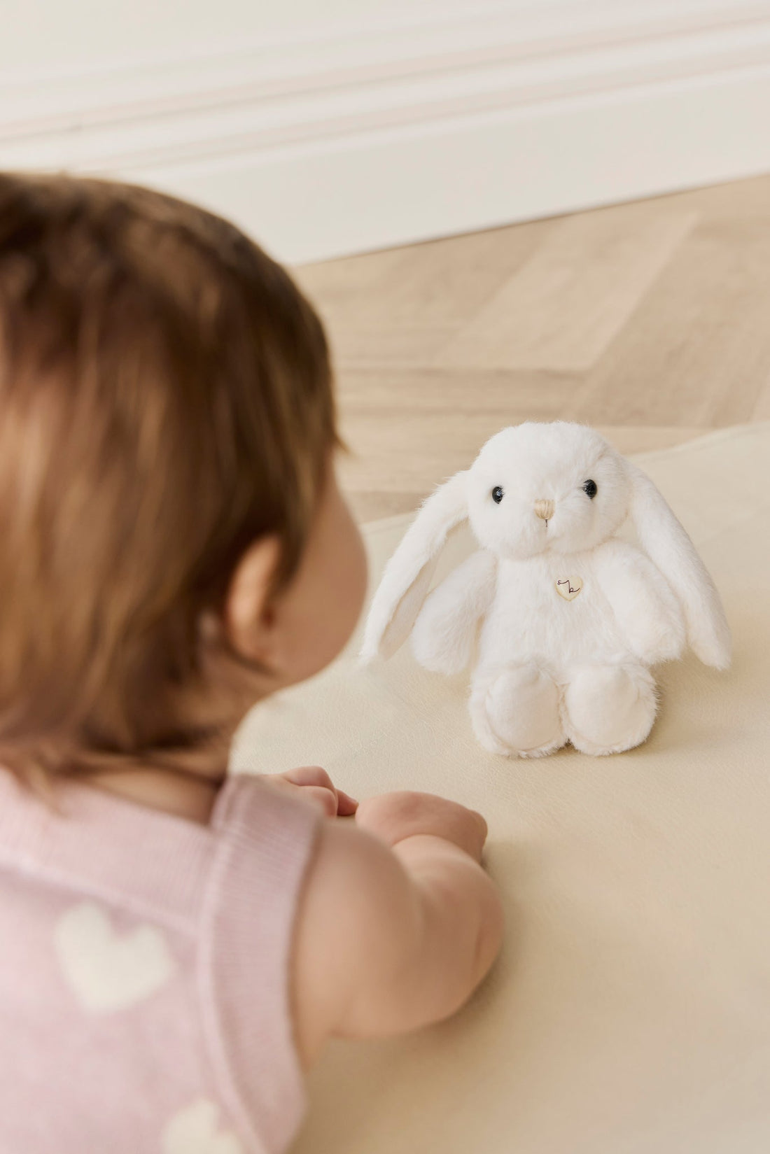 Baby with brown hair and a pink heart-patterned sweater looking at a white stuffed bunny on a light beige surface.