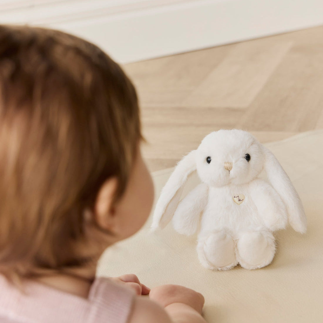 Baby with brown hair and a pink heart-patterned sweater looking at a white stuffed bunny on a light beige surface.