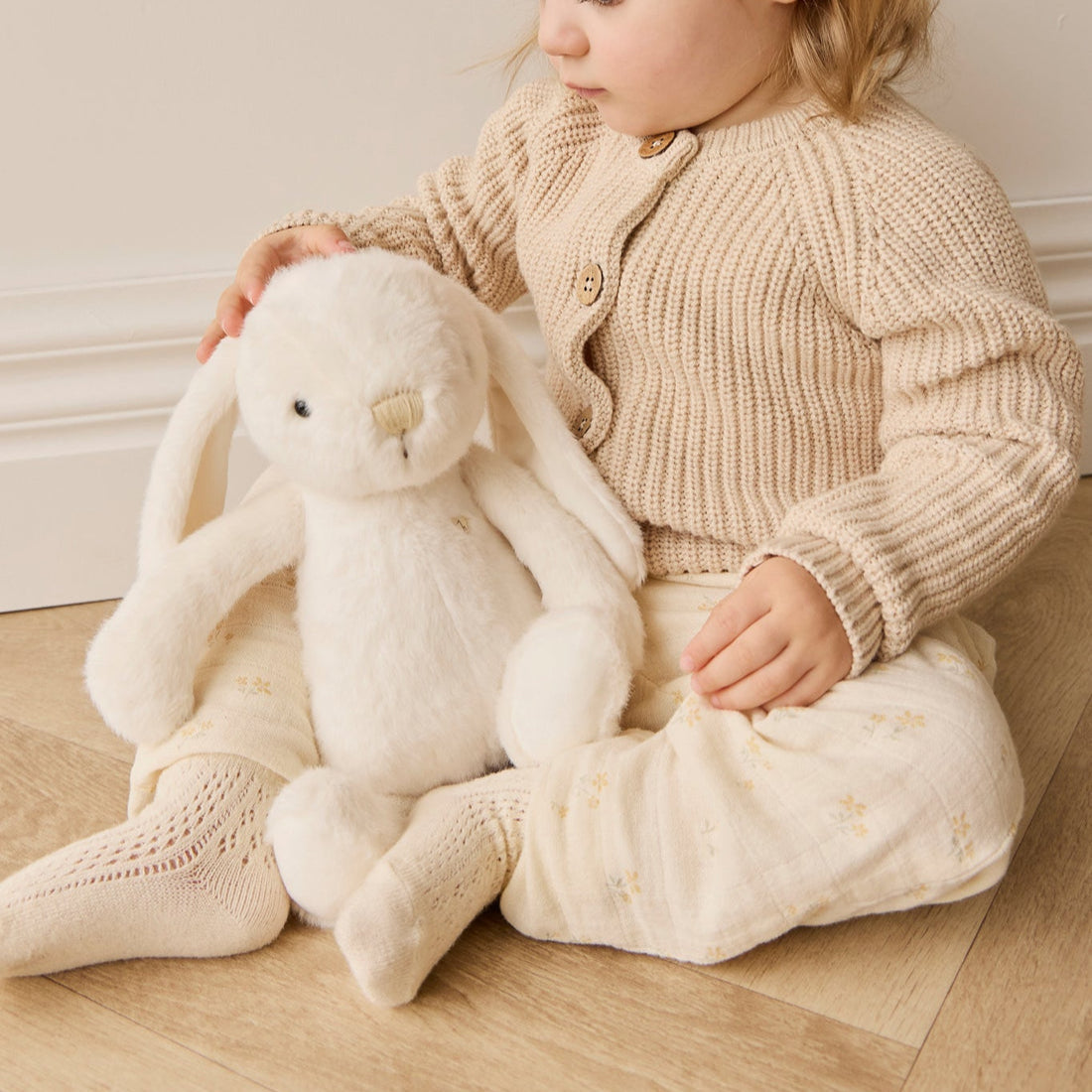 Child sitting with a plush white rabbit, wearing a cream-colored knit cardigan and patterned muslin pants.