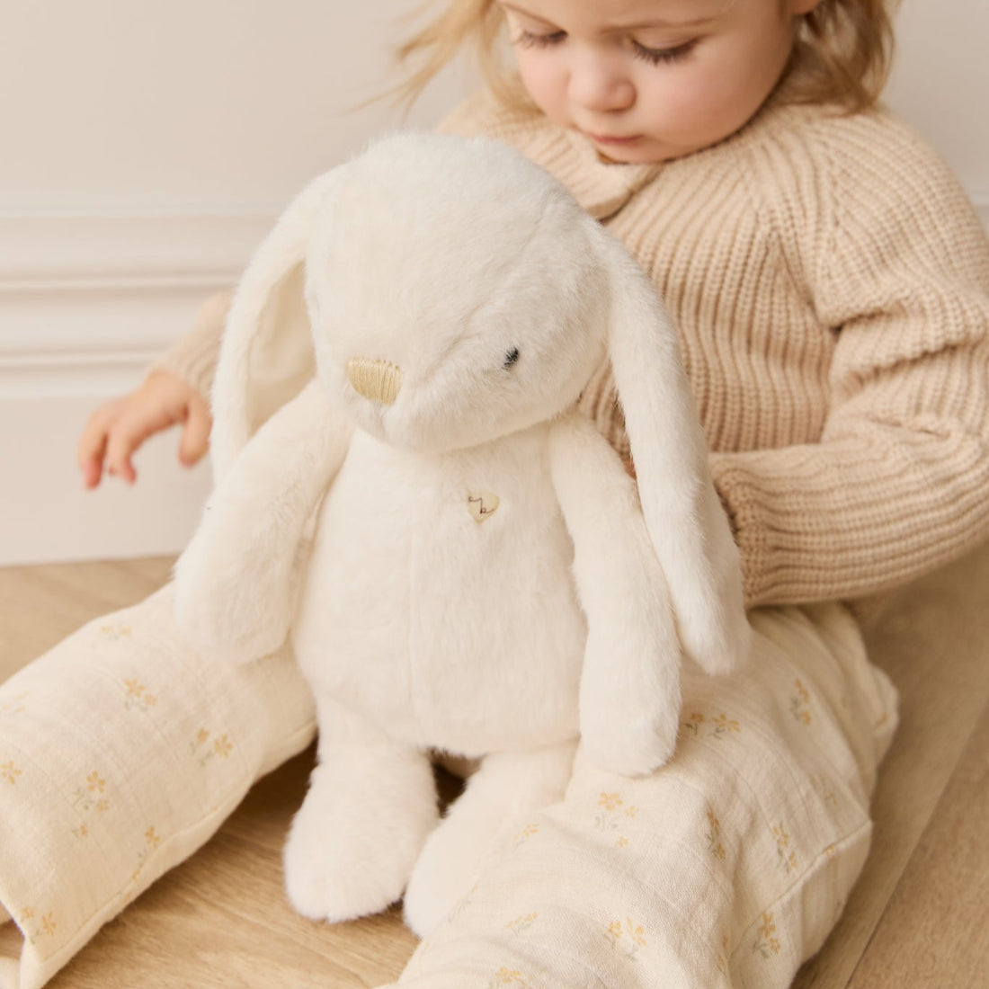 A toddler sits on a wooden floor holding a white plush bunny. The child is wearing a beige sweater and cream-colored patterned pants.