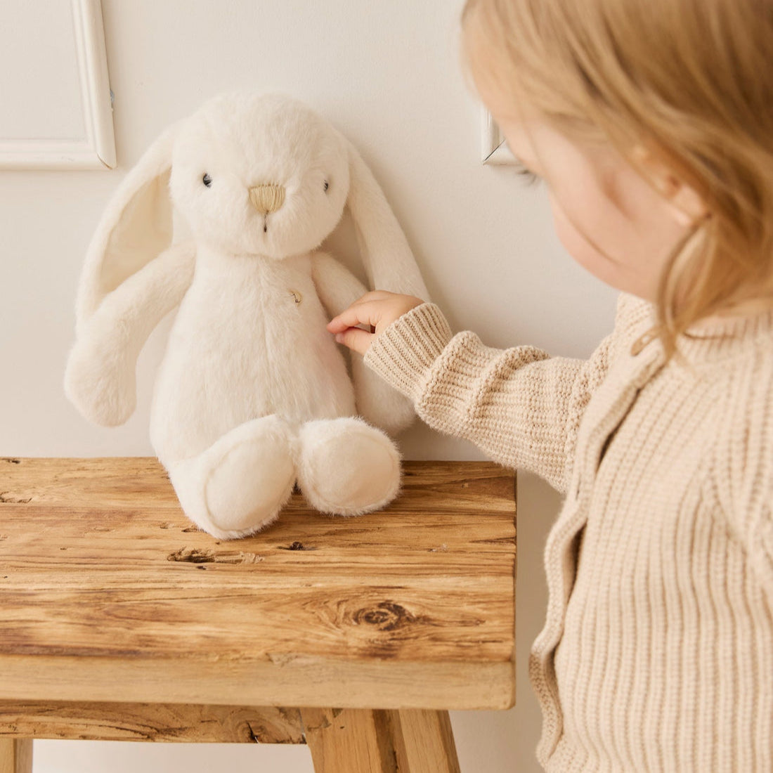 A soft white bunny toy sitting on a wooden table next to a child in a beige sweater.