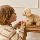 Child in striped sweater and tan overalls facing plush dog toy on wooden table.