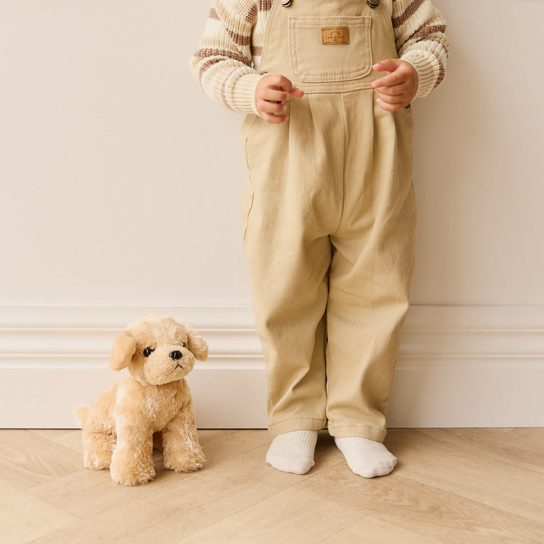 A child in tan overalls and a striped sweater stands beside a stuffed golden retriever toy.