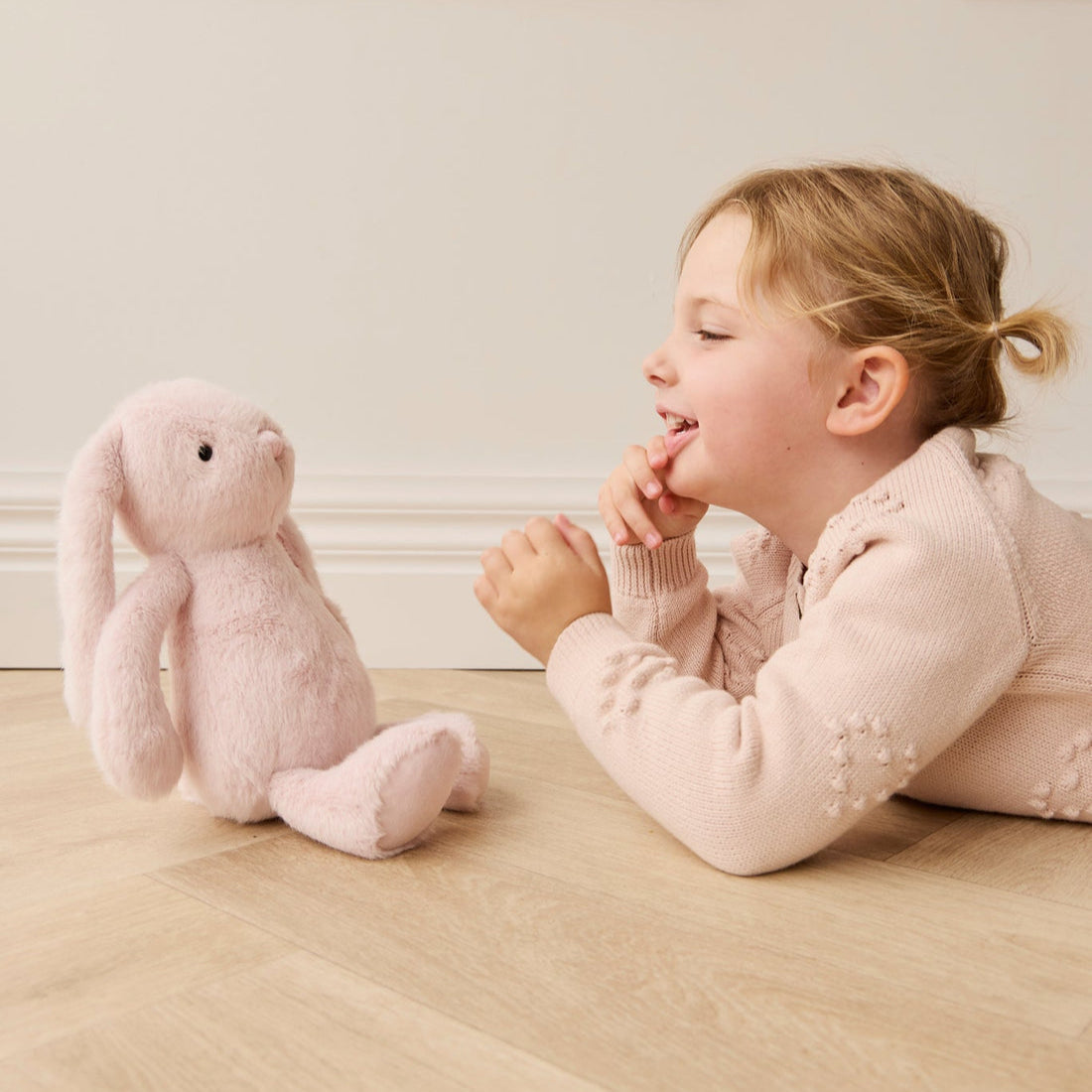A young girl in a pink sweater lying on the floor, facing a pink stuffed bunny.