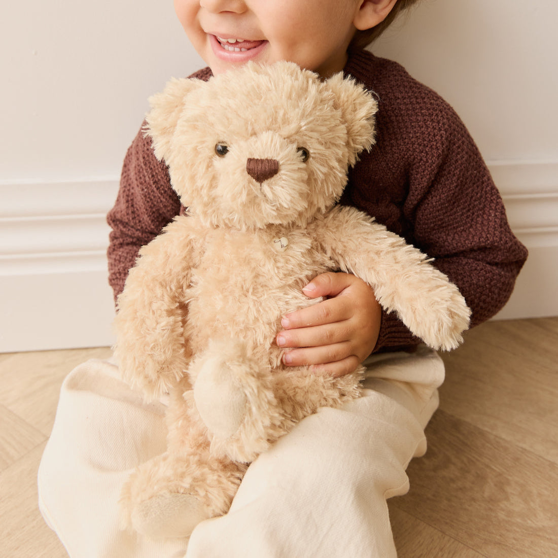 Child hugging a tan plush teddy bear wearing a maroon sweater and off-white pants.
