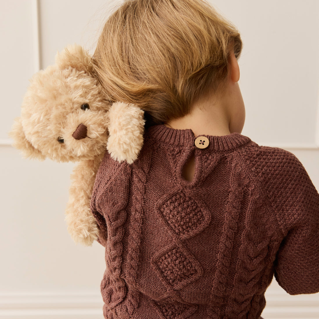 A child with a teddy bear wearing a knitted auburn sweater.