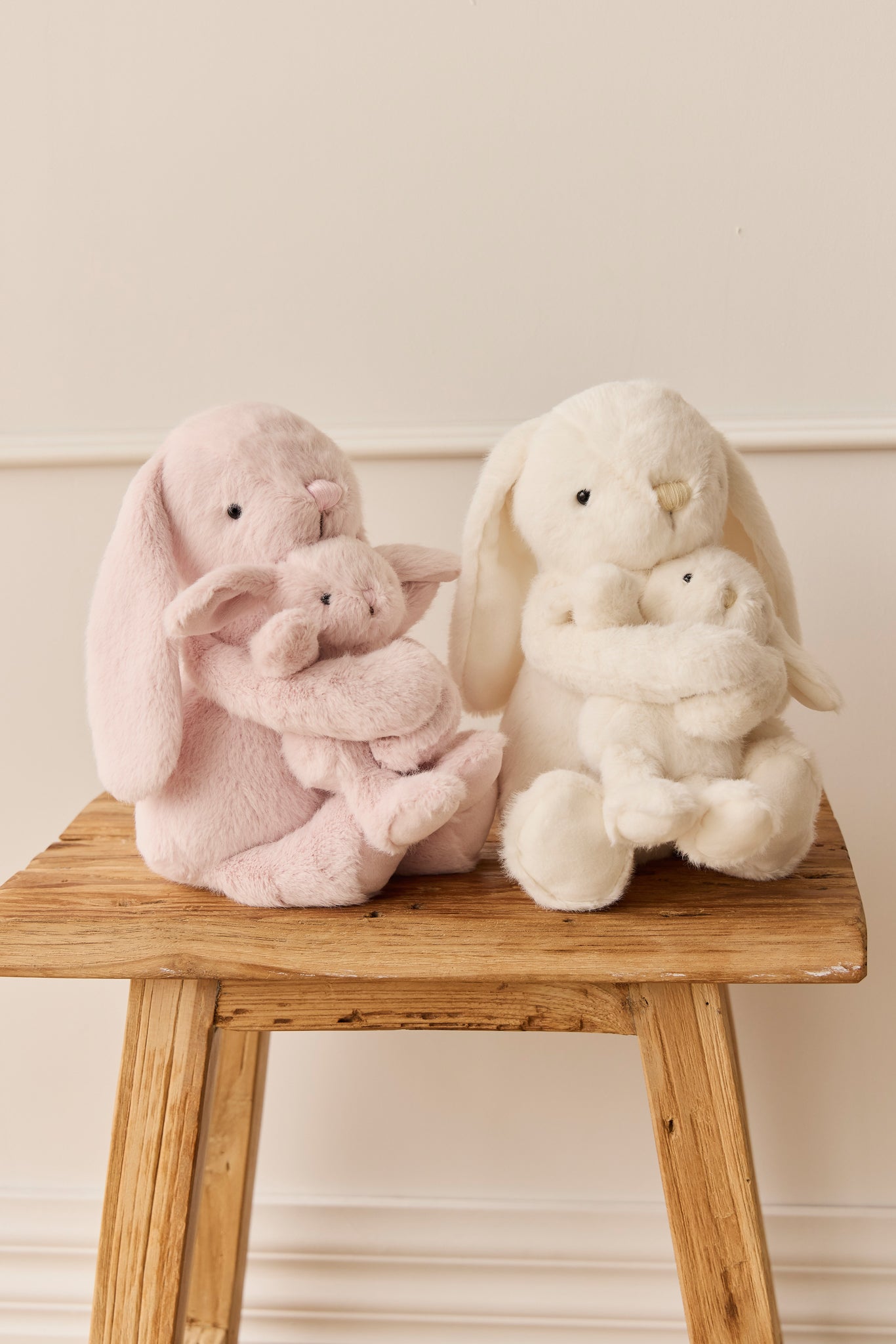 Two plush bunnies, one pink and one white, hugging smaller versions of themselves, sitting on a weathered wooden stool.