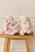 Two plush bunnies, one pink and one white, hugging smaller versions of themselves, sitting on a weathered wooden stool.