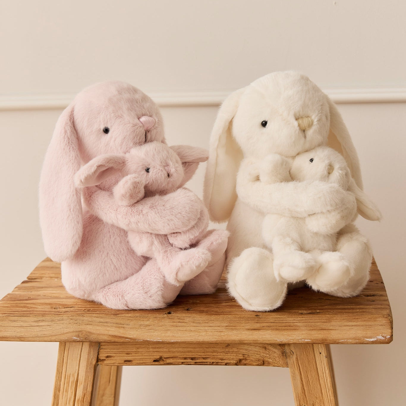 Two plush bunnies, one pink and one white, hugging smaller versions of themselves, sitting on a weathered wooden stool.