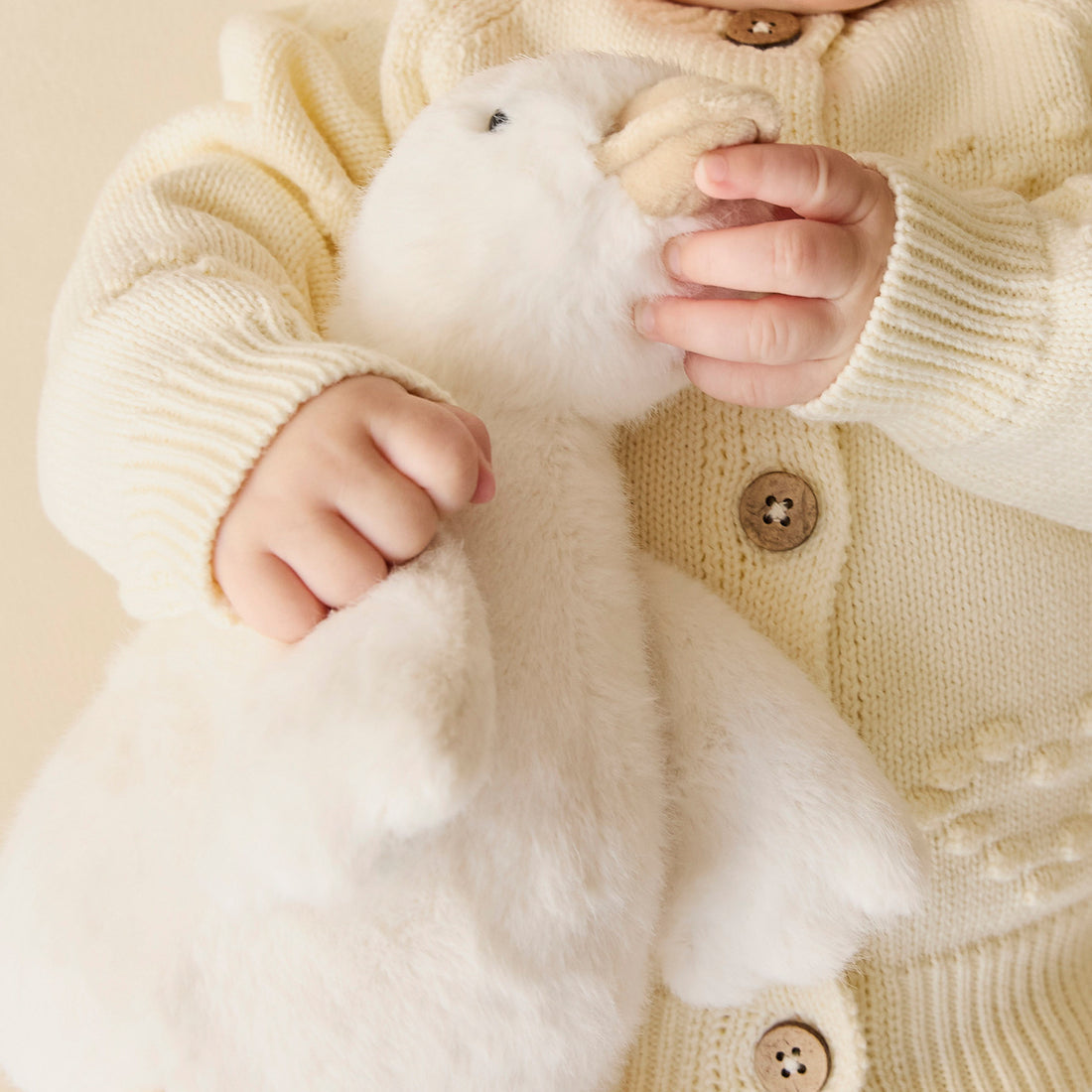 Baby holding a white plush duck while wearing a cream-colored knit cardigan.