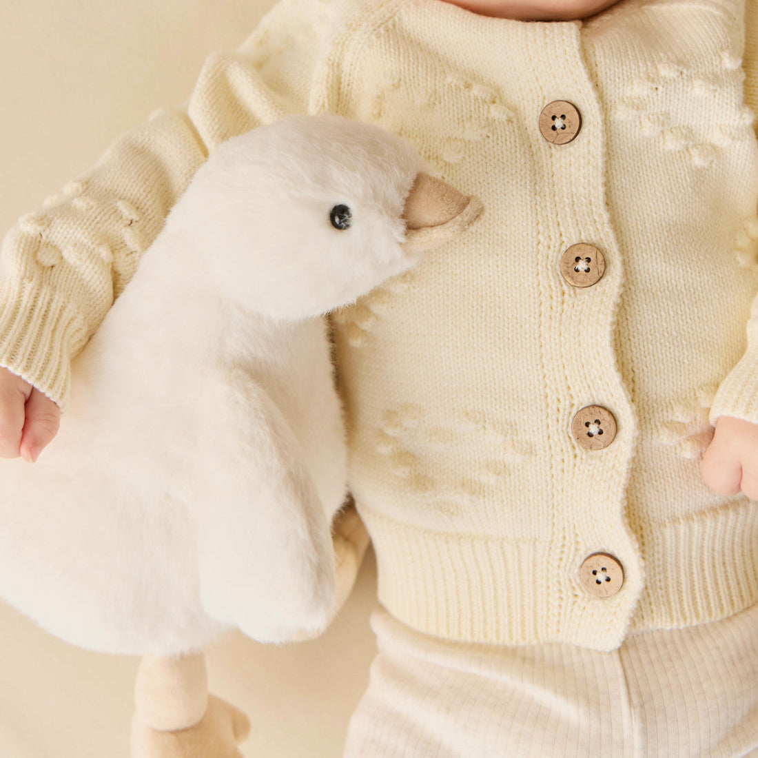 Baby in cream knit cardigan and leggings with plush toy goose.