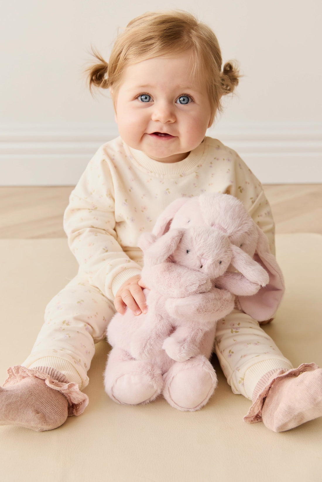 A baby in a floral outfit sits with a plush pink bunny.